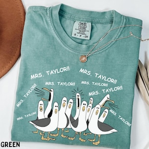 May include: Light green Comfort Colors t-shirt with a graphic of cartoon seagulls and the text "MRS. TAYLOR!!". The shirt has a crew neck and short sleeves. A gold necklace with a pendant is draped on the shirt.