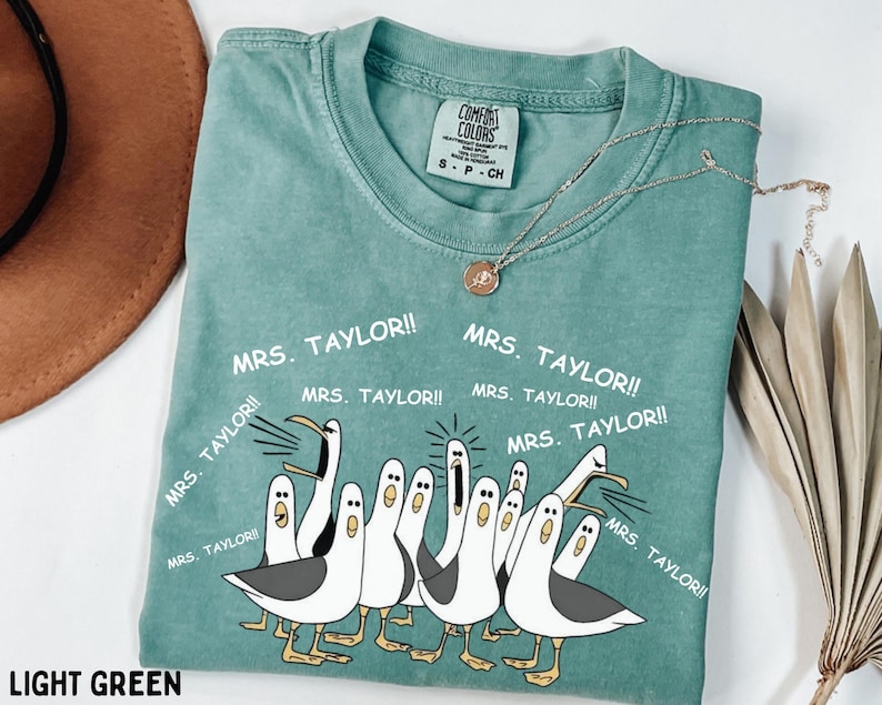 May include: Light green Comfort Colors t-shirt with a graphic of cartoon seagulls and the text "MRS. TAYLOR!!". The shirt has a crew neck and short sleeves. A gold necklace with a pendant is draped on the shirt.