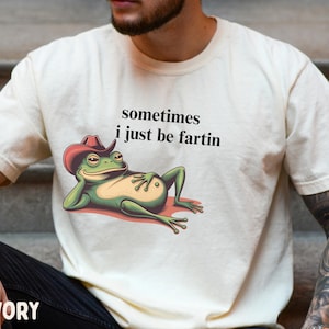 May include: An ivory short-sleeve t-shirt featuring a graphic of a green frog wearing a red cowboy hat, reclining on its side. The text above the frog reads, sometimes i just be fartin, printed in a simple black sans-serif font on the chest.