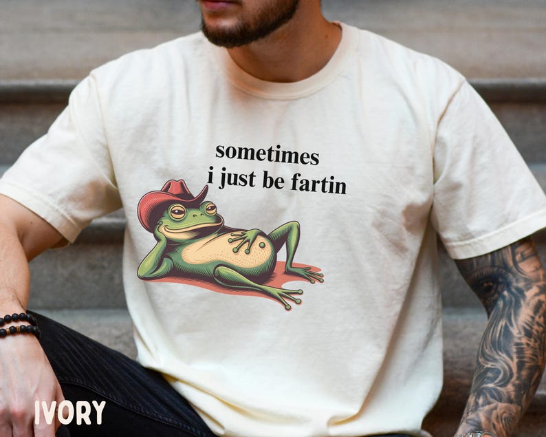 May include: An ivory short-sleeve t-shirt featuring a graphic of a green frog wearing a red cowboy hat, reclining on its side. The text above the frog reads, sometimes i just be fartin, printed in a simple black sans-serif font on the chest.
