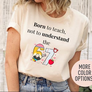 May include: A cream-colored t-shirt with the text "Born to teach, not to understand the 67." The graphic includes a pencil, open book, apple, and alarm clock. The shirt is paired with blue jeans. A pink circle with "MORE COLOR OPTIONS!" is in the bottom right.