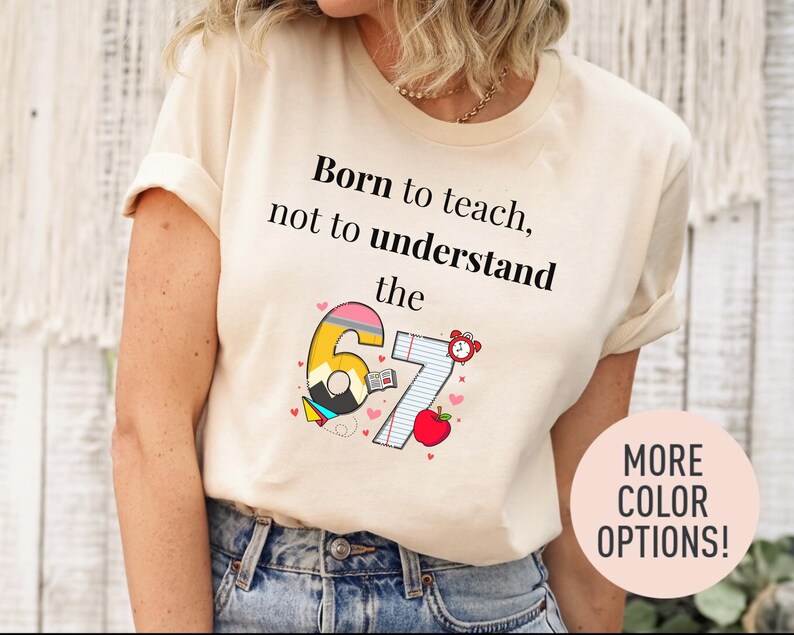 May include: A cream-colored t-shirt with the text "Born to teach, not to understand the 67." The graphic includes a pencil, open book, apple, and alarm clock. The shirt is paired with blue jeans. A pink circle with "MORE COLOR OPTIONS!" is in the bottom right.