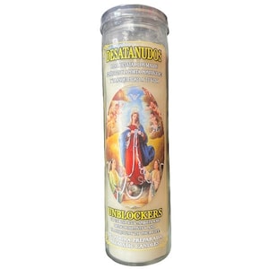 May include: A tall, clear glass candle with a cream-colored label featuring an illustration of a religious figure in red and blue robes. The label includes the text "DESATANUDOS" and "UNBLOCKERS" in gold lettering, along with additional text in Spanish.