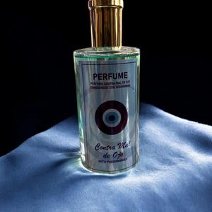 May include: A clear glass perfume bottle with a gold cap. The label features the word "PERFUME" and text in Spanish. The bottle has a blue and purple eye symbol and is sitting on a blue fabric.