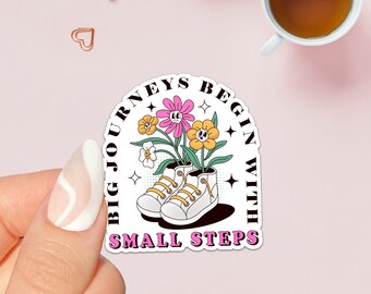 May include: A white sticker with a pink, yellow and green flower design. The flowers are growing out of a pair of white trainers. The text on the sticker reads "Big Journeys Begin With Small Steps".