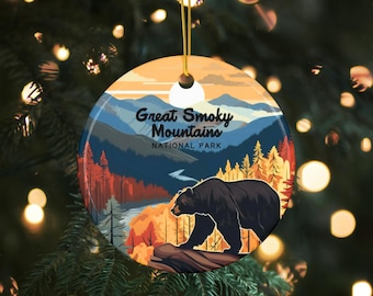 May include: A round ceramic ornament featuring a scenic view of the Great Smoky Mountains National Park. The ornament depicts a black bear walking on a log in a forest setting with a river and mountains in the background. The text "Great Smoky Mountains National Park" is printed on the ornament.