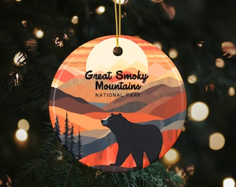 May include: A round ceramic ornament with a sunset scene of the Great Smoky Mountains National Park. The ornament features a black bear walking in the foreground, with a mountain range and trees in the background. The text "Great Smoky Mountains National Park" is printed in black on the ornament.
