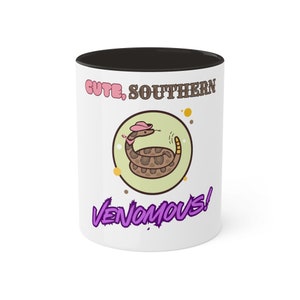 Könnte beinhalten: Weiße Keramiktasse mit schwarzem Rand. Die Tasse zeigt eine Karikatur einer Klapperschlange, die einen rosafarbenen Cowboyhut trägt, und den Text "Cute, Southern Venomous!" aufgedruckt.