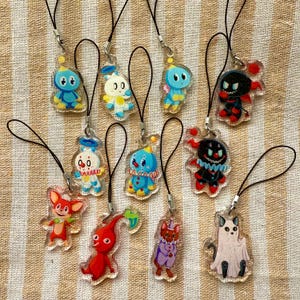 May include: A collection of 11 colorful character charms on black string. The charms are made of clear plastic with painted details. The characters include a clown, a ghost, a jester, a robot, and a few others.
