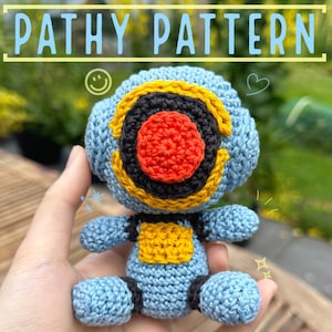 May include: A crocheted blue robot toy with a yellow chest plate and a black and yellow face with a red center. The toy is being held in a hand.