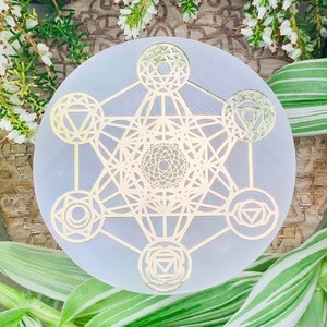 Puede incluir: Una placa de carga de selenita blanca con un diseño geométrico dorado metálico en forma de flor de la vida y siete chakras.