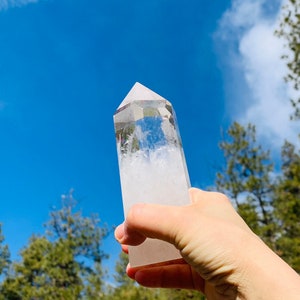 Puede incluir: Una punta de cristal de cuarzo transparente con un interior nublado y blanco. El cristal se sostiene en una mano contra un cielo azul con nubes blancas.