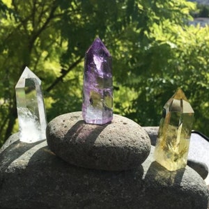 May include: Three crystal points, one clear, one purple, and one yellow, are arranged on top of two gray stones.