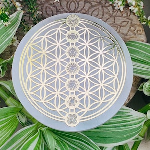 Könnte beinhalten: Ein grauer runder Untersetzer mit einem goldenen Lebensblume-Design und sieben goldenen Chakra-Symbolen in einer vertikalen Linie.
