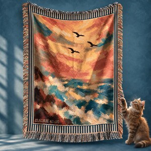 May include: A woven throw blanket featuring a sunset seascape with three birds in flight, crashing waves, and the text Isaiah 41:10. The blanket has a striped border and fringed edges, displaying a palette of orange, yellow, and blue tones.