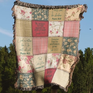 May include: A woven throw blanket featuring a patchwork design of floral, plaid, and solid panels. Several panels contain printed scripture verses including Psalm 16:8, Isaiah 54:17, Psalm 46:5, Luke 12:27, Jeremiah 29:11, and the phrase Be Still and Know.