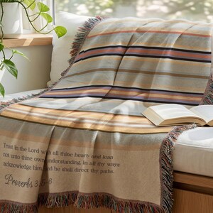 May include: A woven throw blanket featuring horizontal stripes in shades of tan, orange, blue, and cream. The blanket includes fringed edges and the text: Trust in the Lord with all thine heart; and lean not unto thine own understanding. Proverbs 3:5-6.