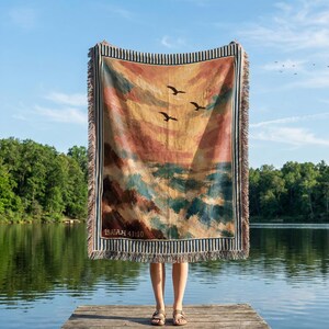 May include: A woven throw blanket featuring a painted landscape of a sunset over a turbulent sea with three birds in flight. The design includes a striped border and the text Isaiah 41:10 printed in the lower left corner of the fabric.