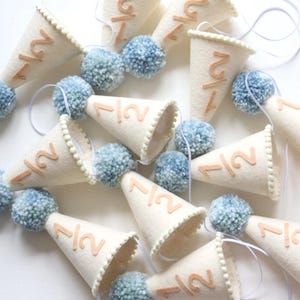 May include: A decorative banner composed of cream-coloured felt cones, each displaying the number "1/2" in a light brown shade. Light blue pom-poms are placed between the cones. The banner is strung on a white string, suitable for celebrations.