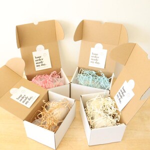May include: Four open cardboard boxes with white interior liners and tissue paper. Each box contains a small white object and a printed tag that reads "Your order made my day."
