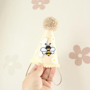 May include: A cream-colored party hat with a tan pom-pom on top. The hat features a white number 1, a cartoon bee, and small white daisies. A brown elastic band is attached for wearing.