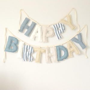 May include: A two-tiered banner with the words "HAPPY BIRTHDAY" in various fabrics and colors. The letters are in blue, white, and beige, with striped and dotted patterns. The banner is suspended by cream-colored ribbons.