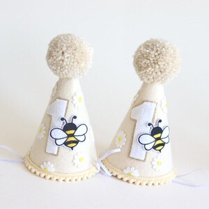 May include: Two cream-colored party hats with a pom-pom on top. Each hat features a white number 1, a bee applique, and daisy accents. The hats have a scalloped trim and white ribbons.