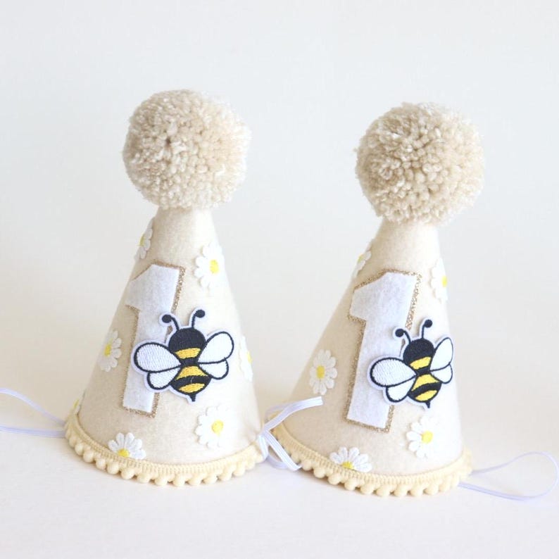 May include: Two cream-colored party hats with a pom-pom on top. Each hat features a white number 1, a bee applique, and daisy accents. The hats have a scalloped trim and white ribbons.