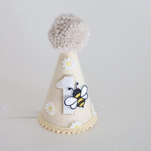 May include: A cream-colored party hat with a beige pom-pom on top. The hat features a number 1, a bee, and daisy accents. The base of the hat has a scalloped trim and a string for securing it.