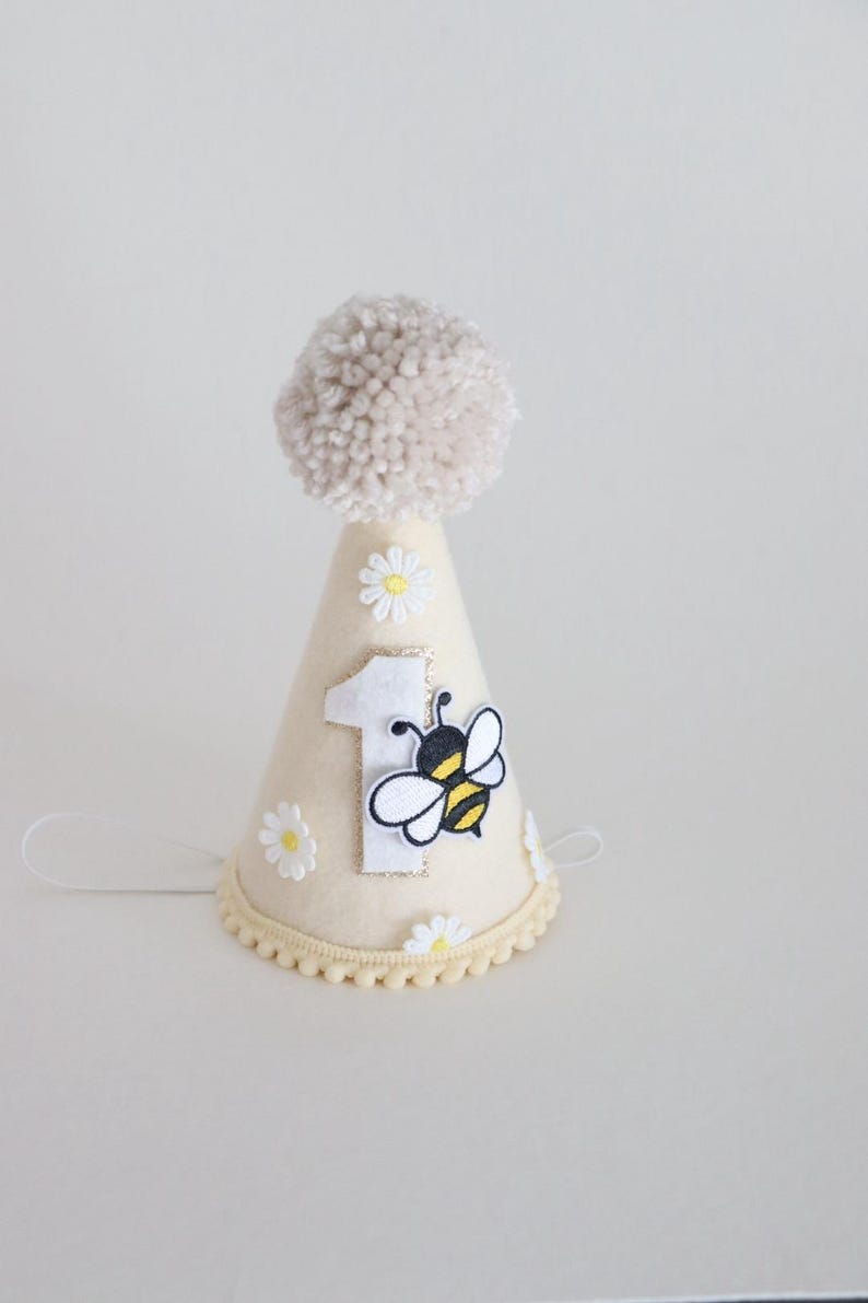 May include: A cream-colored party hat with a beige pom-pom on top. The hat features a number 1, a bee, and daisy accents. The base of the hat has a scalloped trim and a string for securing it.