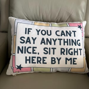 May include: Rectangular white cushion with the text "IF YOU CAN'T SAY ANYTHING NICE, SIT RIGHT HERE BY ME" in navy blue. The cushion has a colourful border with stars.