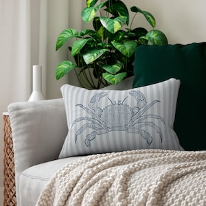 May include: A decorative pillow with a blue crab illustration on a striped background. The pillow is rectangular and rests on a light gray sofa, with a green pillow and a plant in the background. A beige blanket is draped over the sofa.