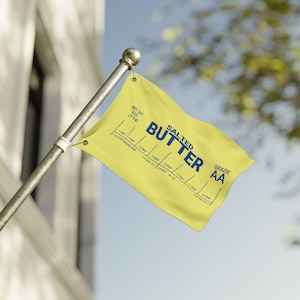 May include: A yellow flag displaying the words "SALTED BUTTER" in blue, with a measurement guide. The flag also features "GRADE AA" and "NET WT. 113g". The flag is attached to a silver pole, against a sky background.