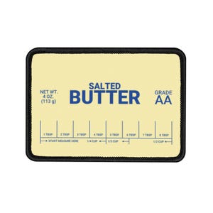 Puede incluir: Un parche rectangular con fondo amarillo claro y borde negro. El texto "SALTED BUTTER" es azul. Incluye medidas en cucharadas y tazas, con un peso neto de 113 g y una calidad AA.
