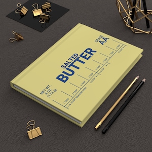 May include: A pale yellow hardcover notebook featuring a blue graphic design that mimics a salted butter wrapper. The cover displays the text "SALTED BUTTER", "GRADE AA", and "NET WT. 4 OZ. (113 g)" along with measurement markings for tablespoons.