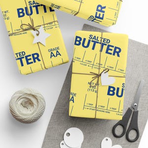 May include: Yellow gift wrap with a blue "SALTED BUTTER" design, measuring guides, and decorative twine. The wrapped gifts are adorned with white star and heart-shaped tags. A ball of twine and scissors are also visible.