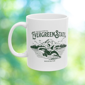 May include: White ceramic mug with a green graphic design featuring a girl riding an orca whale in front of a mountain and trees. The text "Greetings from the Evergreen State Washington" is printed on the mug.