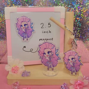 May include: A display of acrylic charms and magnets featuring a cartoon character with pink hair and a microphone. The display includes a 6.35 cm magnet, a keychain, and a stand-up charm. The background is pink and white.