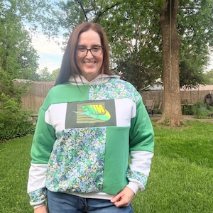 May include: A green, white, and floral patchwork hoodie with the Nike logo. The hoodie features a white hood and sleeves, with a floral pattern on the front and sleeves. The Nike logo is in green and yellow.