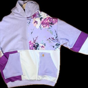 May include: A lavender hoodie with floral print and purple and white accents. The hoodie features a floral pattern on the upper right side and sleeves, with a Carhartt pocket. The design incorporates a mix of solid and patterned fabrics.