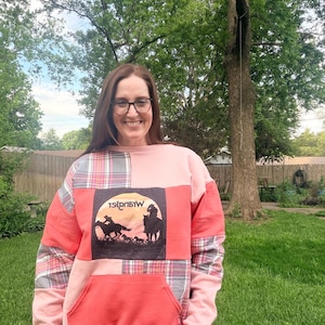 May include: A patchwork sweatshirt in shades of pink and coral, featuring a large front pocket and plaid accents. The center panel displays a black graphic of cowboys and horses with the word "Wrangler" in reverse.