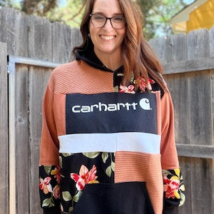 May include: A patchwork hoodie with the Carhartt logo. The hoodie features a mix of colors and patterns, including a rust-colored knit, floral print, and black fabric. The Carhartt logo is displayed in a white rectangle.