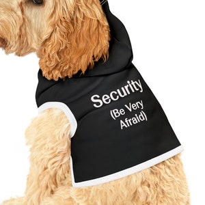 May include: A light brown dog wearing a black hooded vest with white trim. The vest has the text "Security (Be Very Afraid)" printed on it.