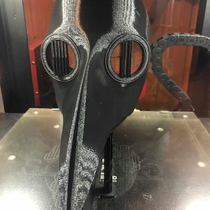 May include: A black 3D printed plague doctor mask with a long beak and two eye holes with metal grates. The mask is sitting on a gray surface.