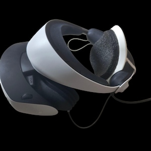 Op de afbeelding: Een wit en zwart virtual reality-headset met een zwart hoofdband en een zwart snoer. De headset heeft een zwarte knop met de tekst "AUDIO".