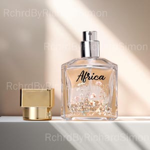 May include: A clear, square perfume bottle with a silver sprayer and a gold cap. The bottle contains a light amber liquid and features the word "Africa" in black script, surrounded by a floral design.