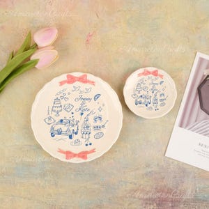 May include: Two off-white ceramic plates with scalloped edges. The plates feature a blue illustration of a car, cake, and text that reads "Jonny & Kate". Pink bows are at the top and bottom of the larger plate and top of the smaller plate.