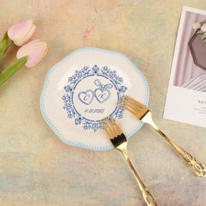 Könnte beinhalten: Ein weißer Teller mit blauem Blumendesign und zwei goldenen Gabeln. Der Teller hat einen blauen Rand und ein blaues Blumendesign mit zwei Herzen und dem Datum 11.12.2025. Rosa Tulpen sind im Hintergrund.