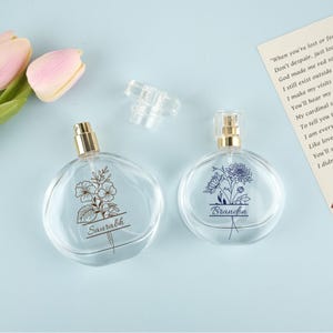 Puede incluir: Dos frascos de perfume de vidrio transparente con pulverizadores dorados y diseños florales. Una botella tiene el nombre "Saurabh" y la otra "Brandon" impresos. Las botellas están sobre una superficie azul claro.