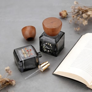 Può includere: Due flaconi di profumo in vetro nero con coperchi in legno e disegni floreali. Un flacone ha un vaporizzatore color oro. I flaconi sono su una superficie grigia accanto a un libro aperto e fiori secchi.